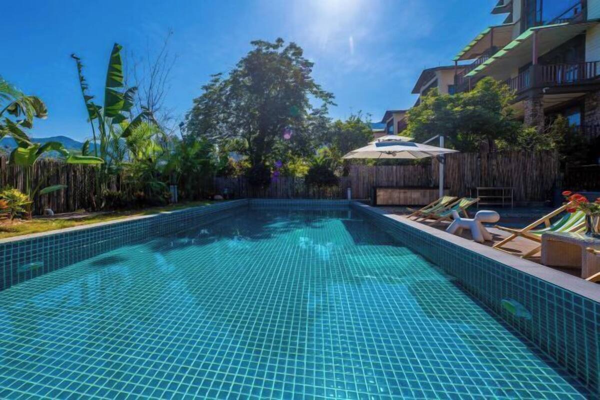 A swimming pool with tiled flooring, surrounded by lounge chairs and umbrellas, with a wooden fence and tropical plants.
