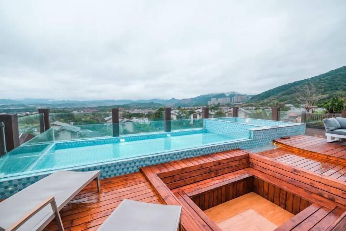 A rooftop pool area with a glass balustrade, wooden deck, and a view of the city and hills in the background.