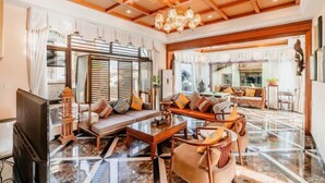 Amplia sala de estar con un sofá grande, una mesa de centro y un televisor. La habitación cuenta con techo de madera, un candelabro y un ventanal con cortinas.