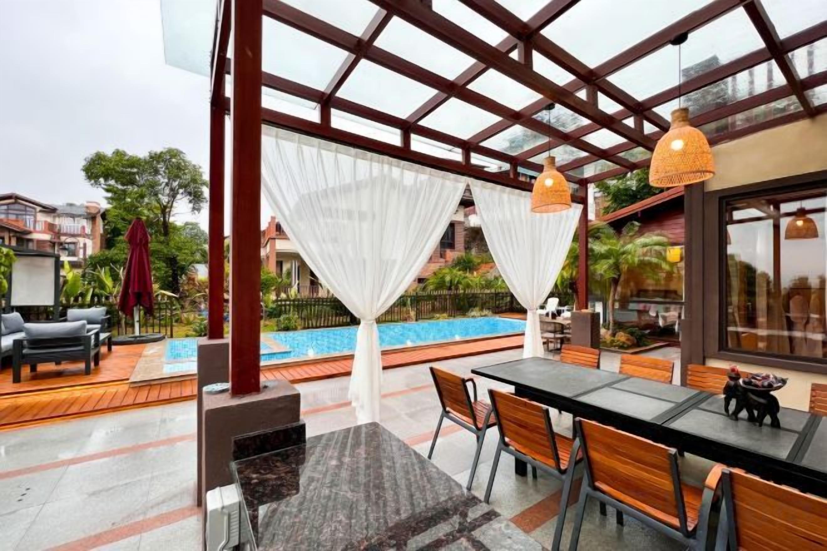 An outdoor seating area with a pool view, covered by a glass structure, and furnished with tables and chairs.