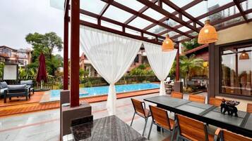 An outdoor seating area with a pool view, covered by a glass structure, and furnished with tables and chairs.