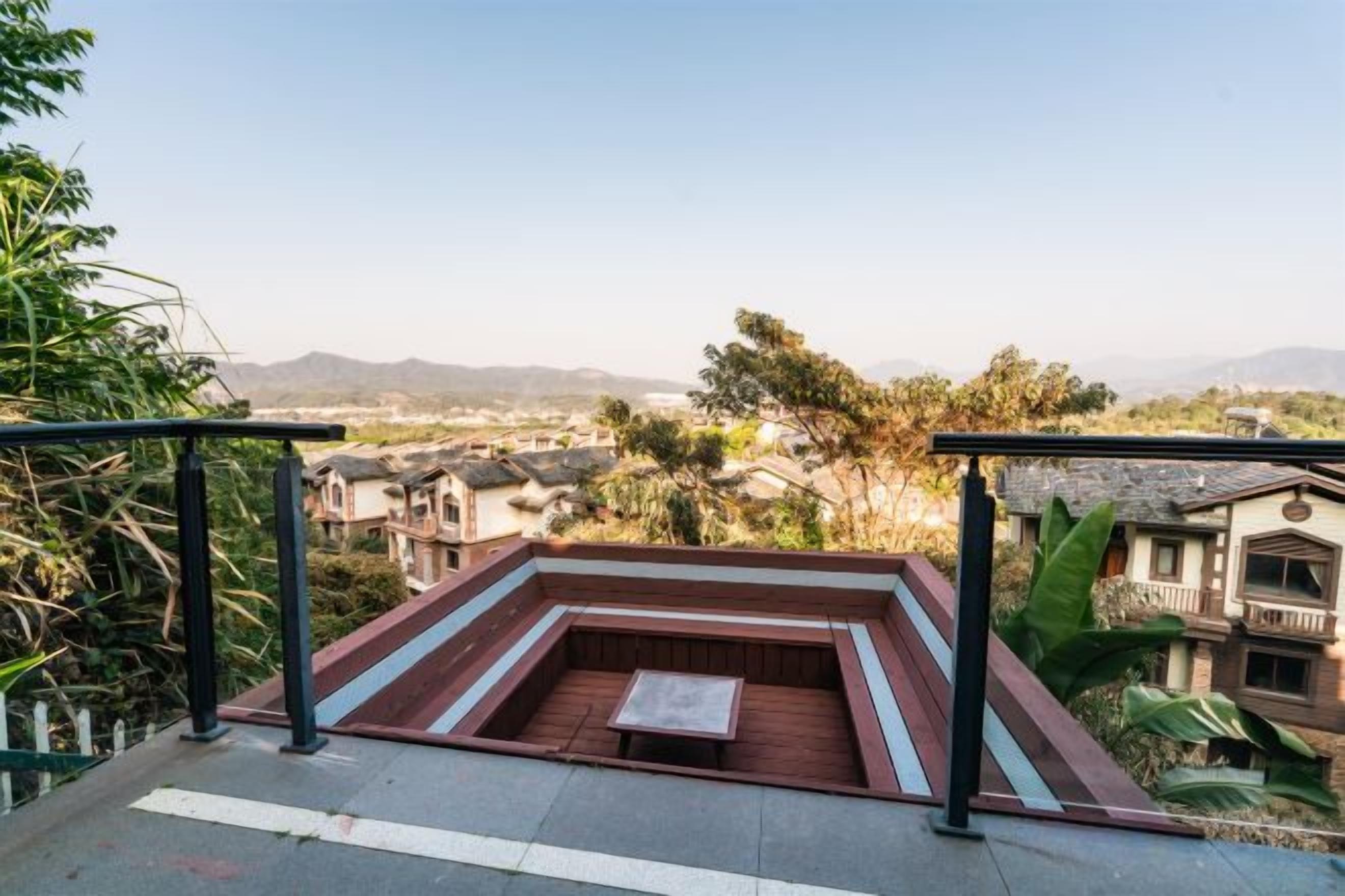 A rooftop terrace with a seating area, surrounded by greenery and overlooking a residential area with houses and mountains in the distance.