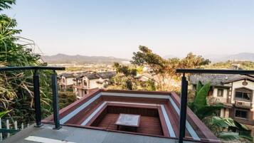 A rooftop terrace with a seating area, surrounded by greenery and overlooking a residential area with houses and mountains in the distance.