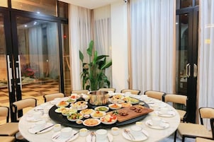 Una mesa de comedor redonda con varios platos de comida, rodeada de sillas, en una habitación con ventanas grandes y cortinas.