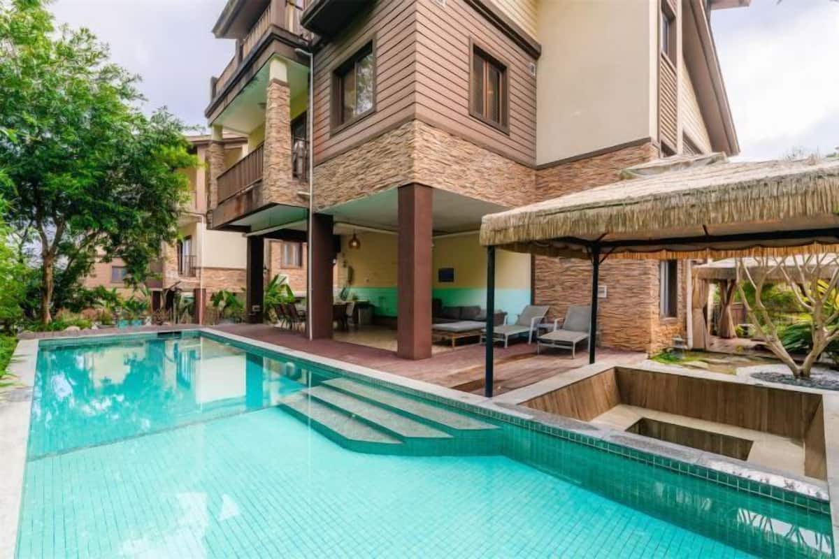 A pool area with a clear blue pool, a covered seating area with chairs, and a building with a stone facade.