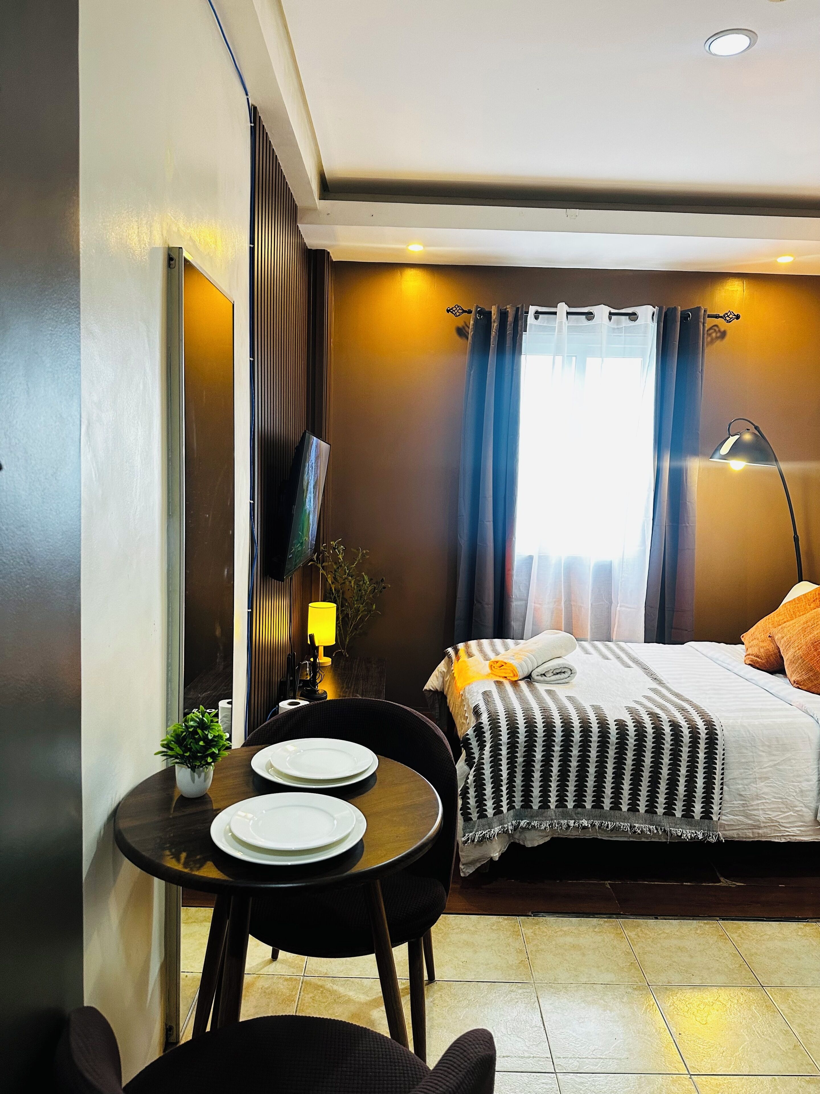 A hotel room with a bed, a desk with plates, a chair, a lamp, and a window with curtains.