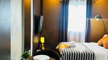 A hotel room with a bed, a desk with plates, a chair, a lamp, and a window with curtains.