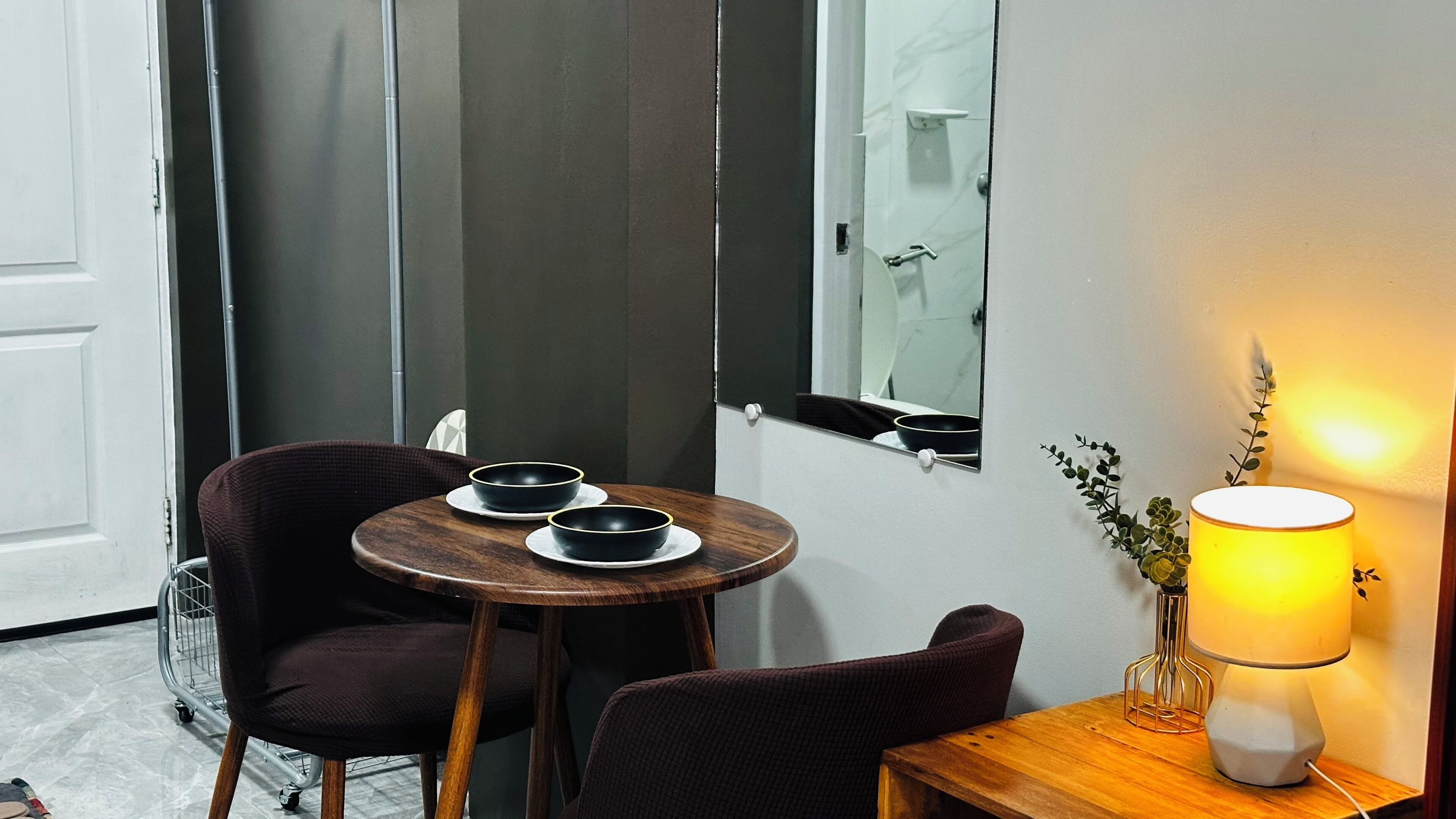 A room with a wooden table, two chairs, a mirror, a lamp, and a door.