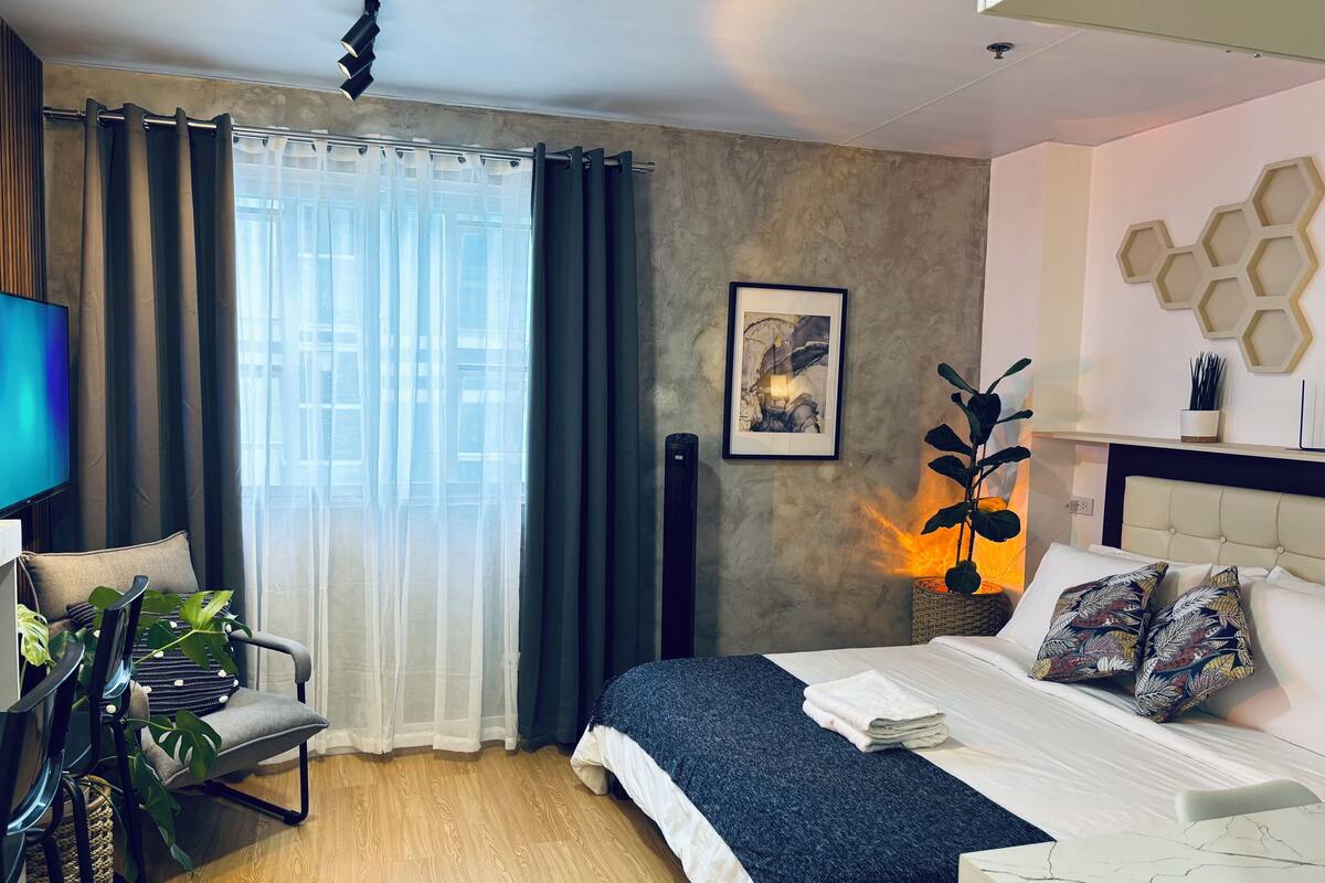 A modern hotel room with a bed, a desk, and a chair. There is a window with curtains, a wall-mounted artwork, and a decorative wall piece.