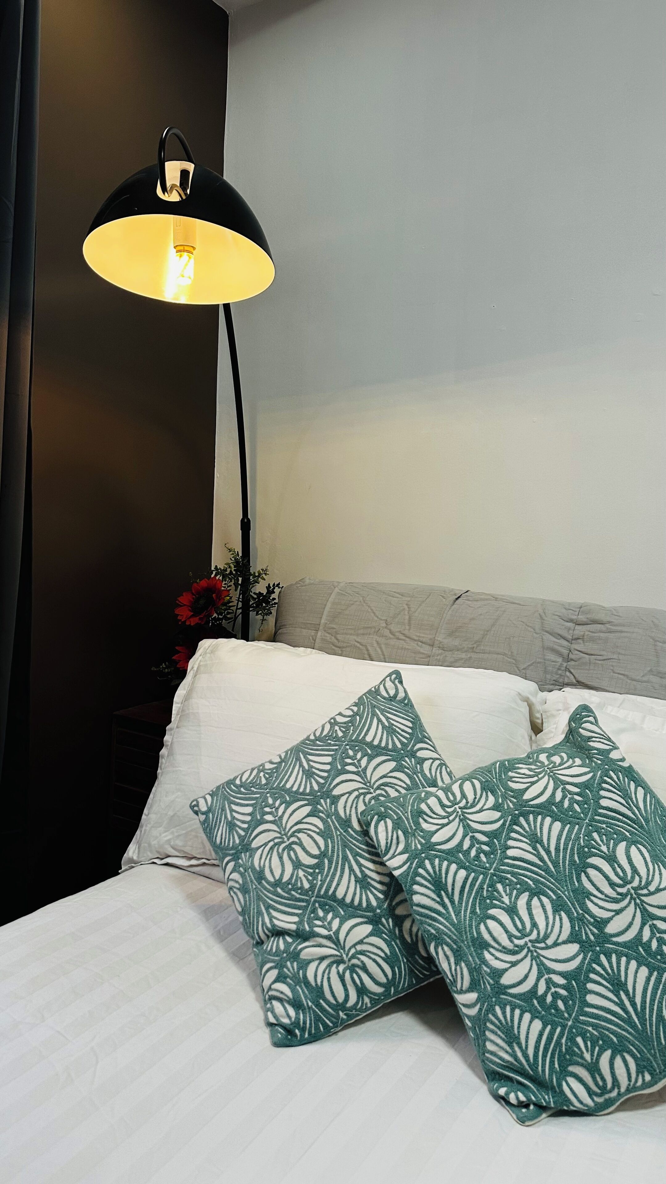 A bed with white bedding and green patterned pillows, a black floor lamp, and a wall with a dark panel.