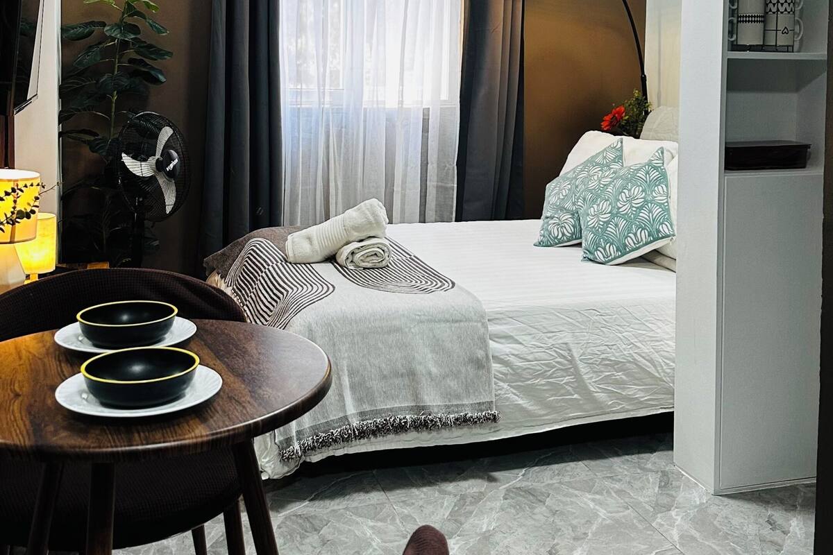 A compact hotel room with a bed, a desk with a chair, a small table with bowls, a potted plant, a lamp, and a window with curtains.