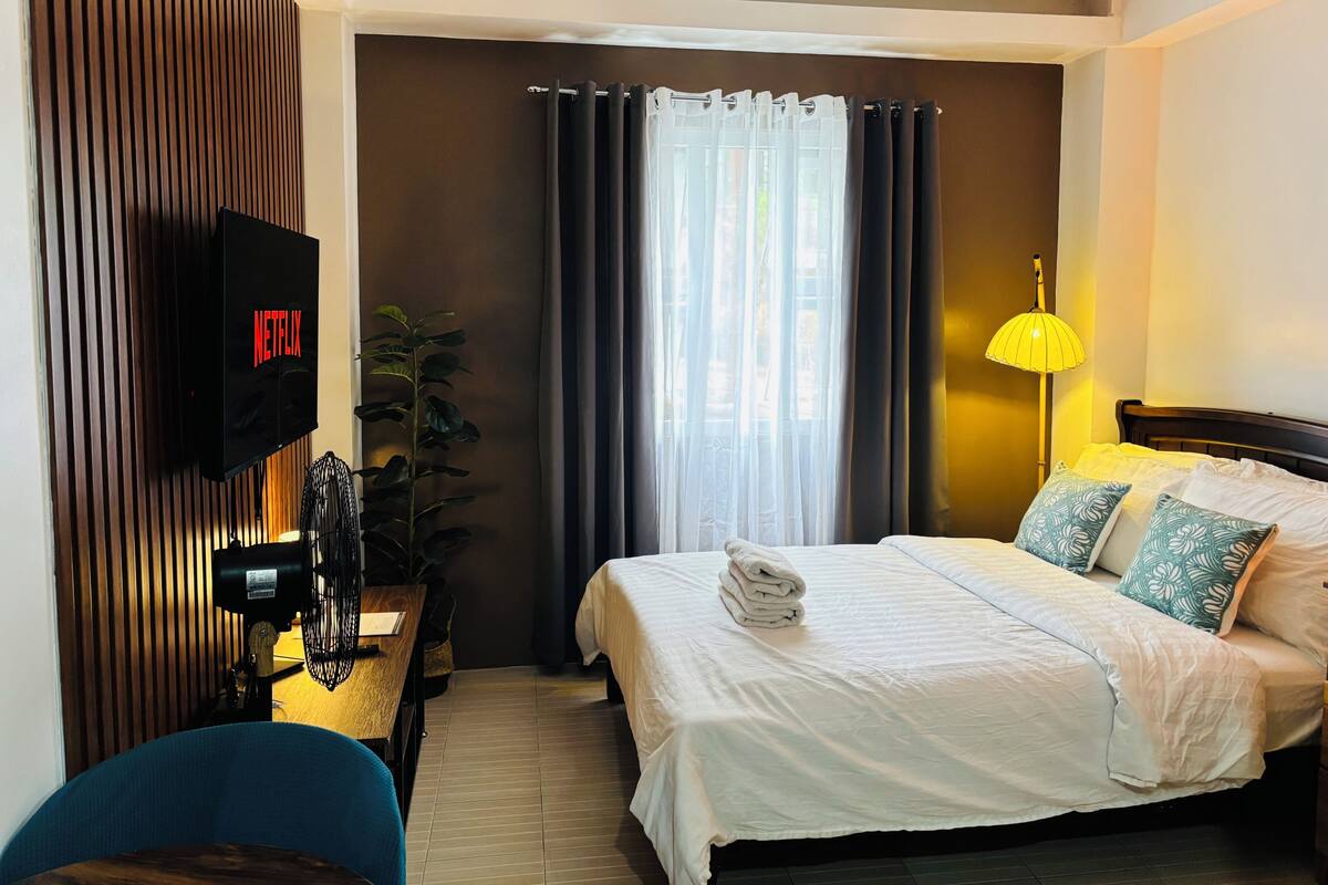 A hotel room with a bed, a desk with a lamp, a TV mounted on the wall, and a potted plant.