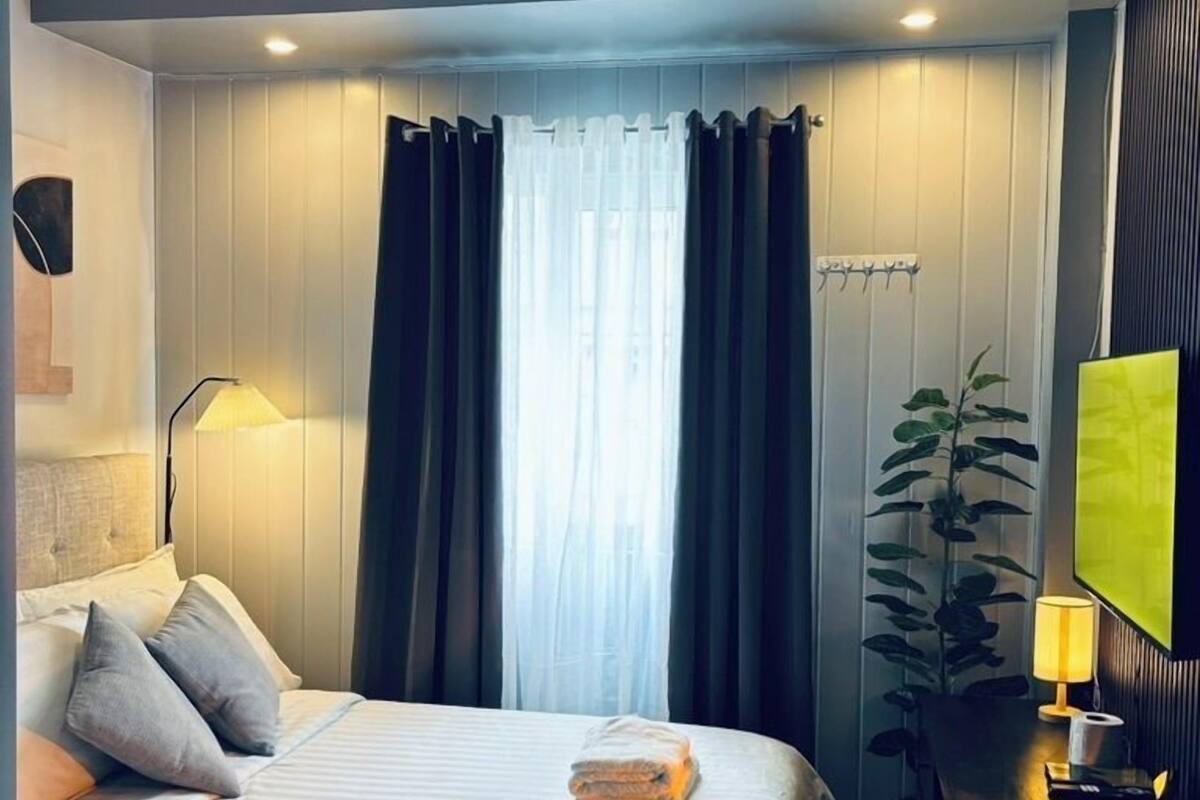 A modern hotel room with a neatly made bed, a desk with a lamp, and a large window with curtains.