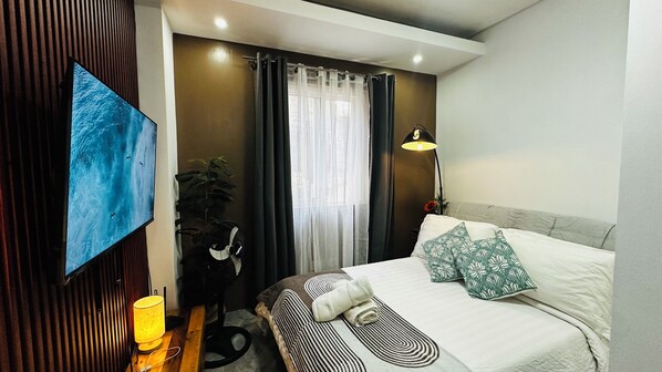 A modern hotel room with a bed, a desk with a lamp, a large window with curtains, and a wall-mounted TV. - Smart Stay 2600 (Baguio)