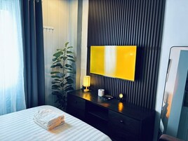 Une chambre d’hôtel avec un lit, une table de chevet avec une lampe, un téléviseur à écran plat fixé à un mur sombre et une plante en pot.