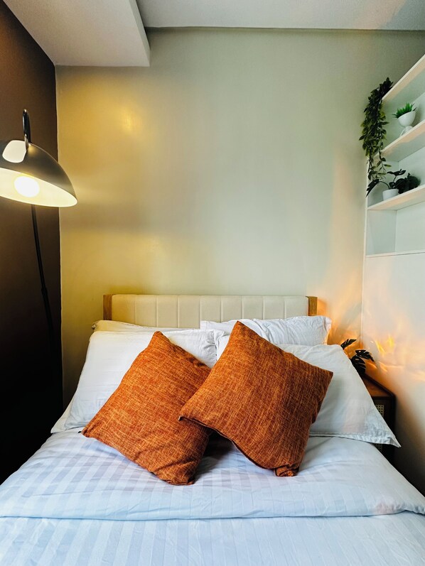 Una cama con sábanas blancas y dos almohadas naranjas, una lámpara de noche y un estante fijado a la pared con plantas.