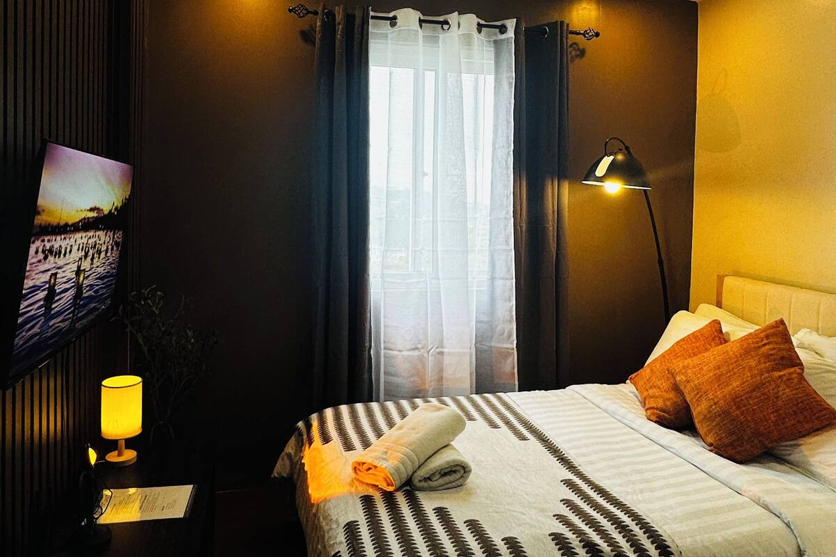 A hotel room with a bed, a TV mounted on the wall, a lamp, and a window with curtains.