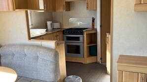 A compact RV kitchen with wooden cabinets, a built-in oven, and a small dining area with a cushioned chair. - Home by the Sea - Adventure Village Access (Great Yarmouth)