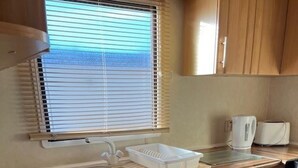 A compact kitchen with wooden cabinets, a sink, and a window with blinds. - Home by the Sea - Adventure Village Access (Great Yarmouth)