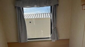 A small, single-room accommodation with two beds, a window with curtains, and a view of a building outside. - Home by the Sea - Adventure Village Access (Great Yarmouth)