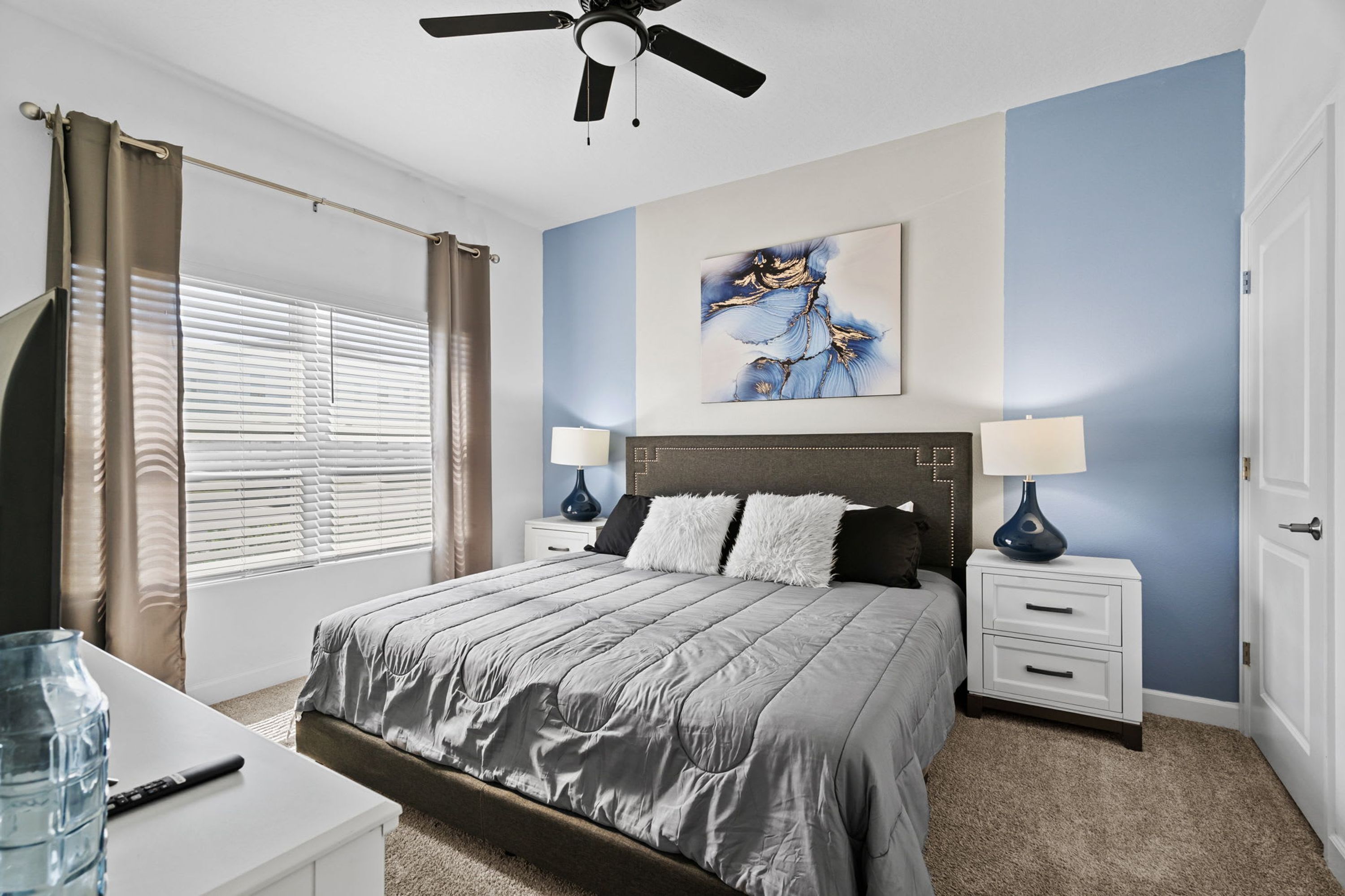 Une chambre à coucher comprenant un lit, deux tables de chevet avec des lampes, un ventilateur de plafond, une grande fenêtre avec des rideaux et un tableau mural.