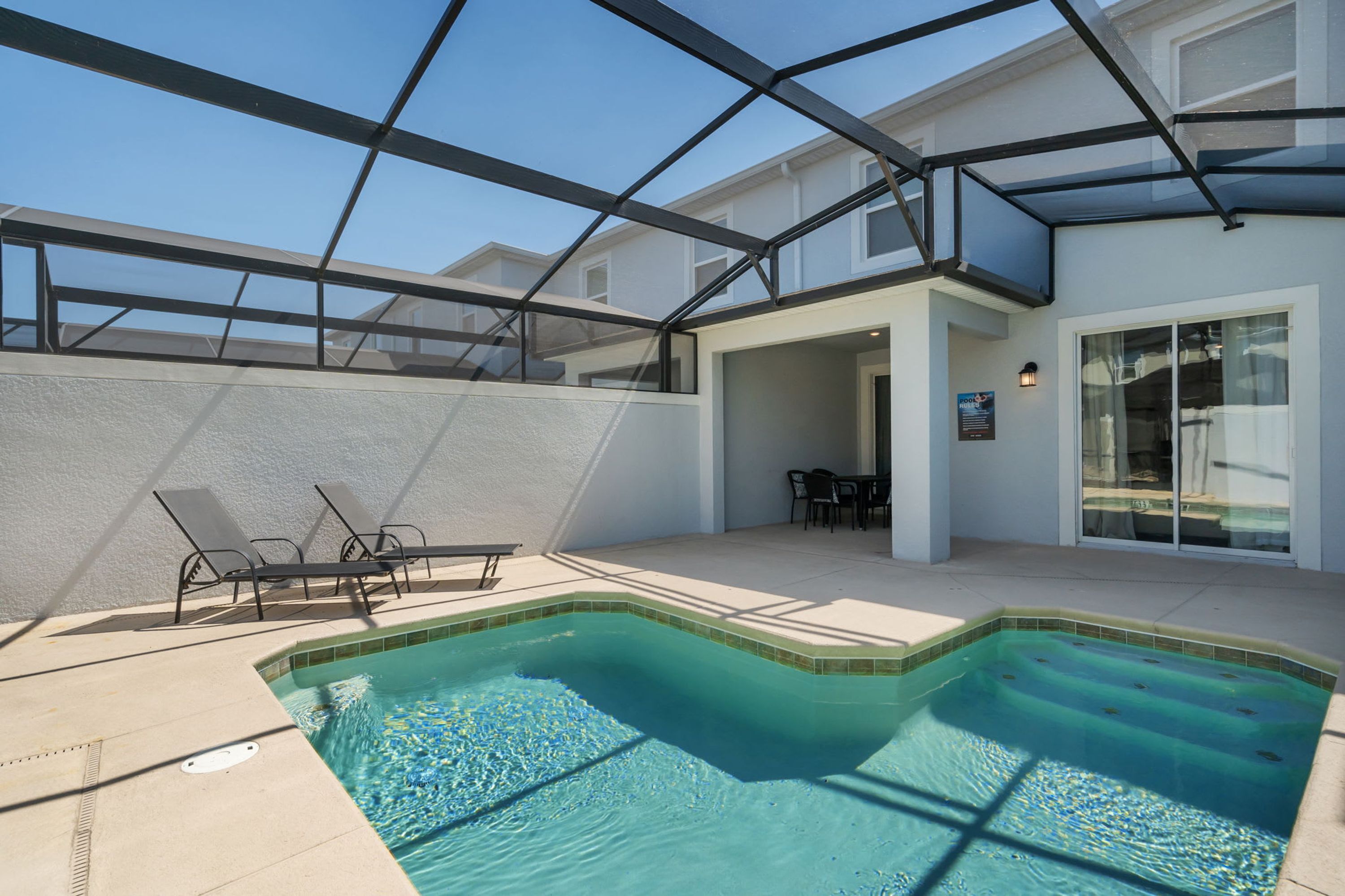 A swimming pool with a glass enclosure, lounge chairs, and a patio area.
