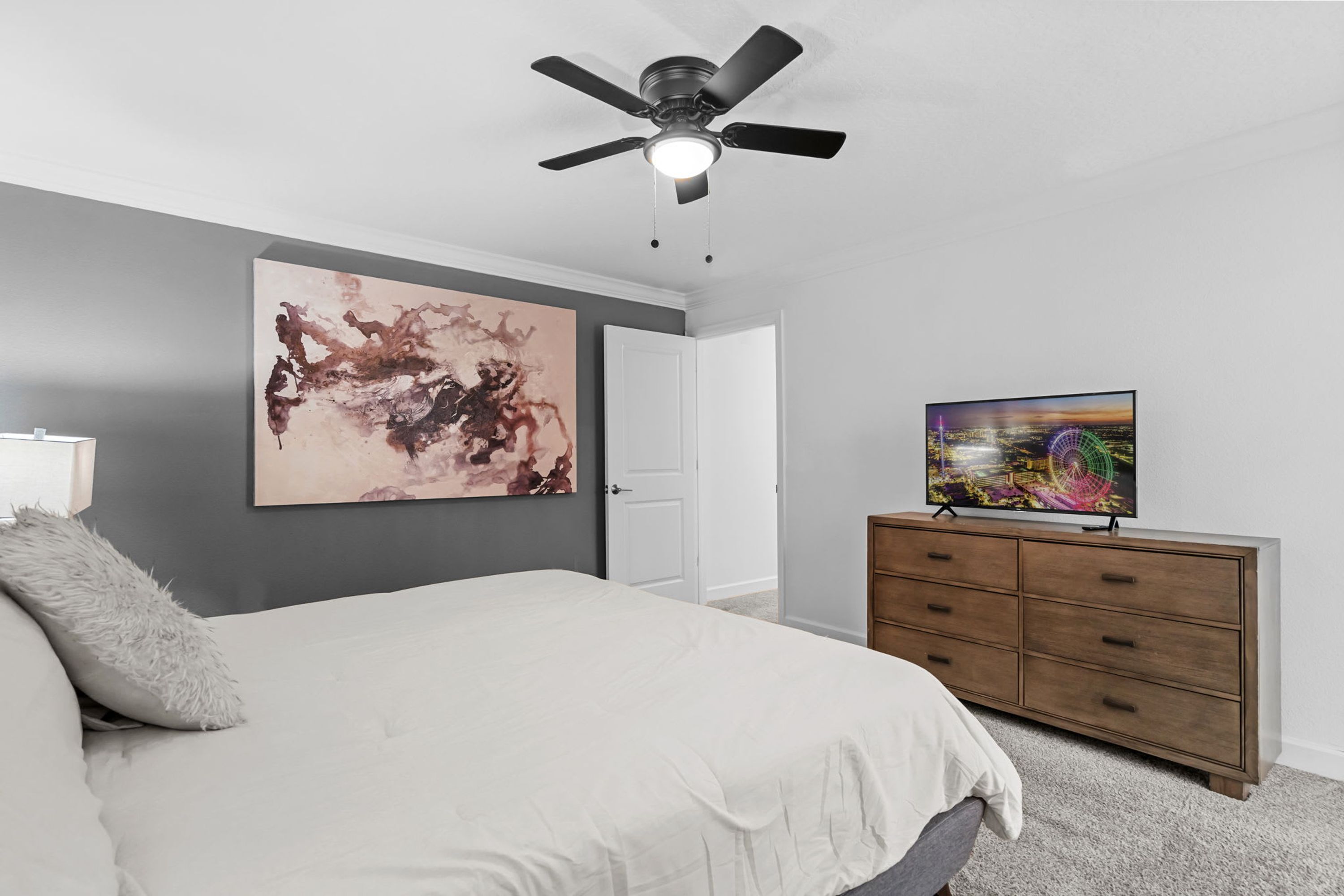 Een slaapkamer met een bed, een plafondventilator, een groot abstract schilderij, een televisie op een dressoir, en een deur.