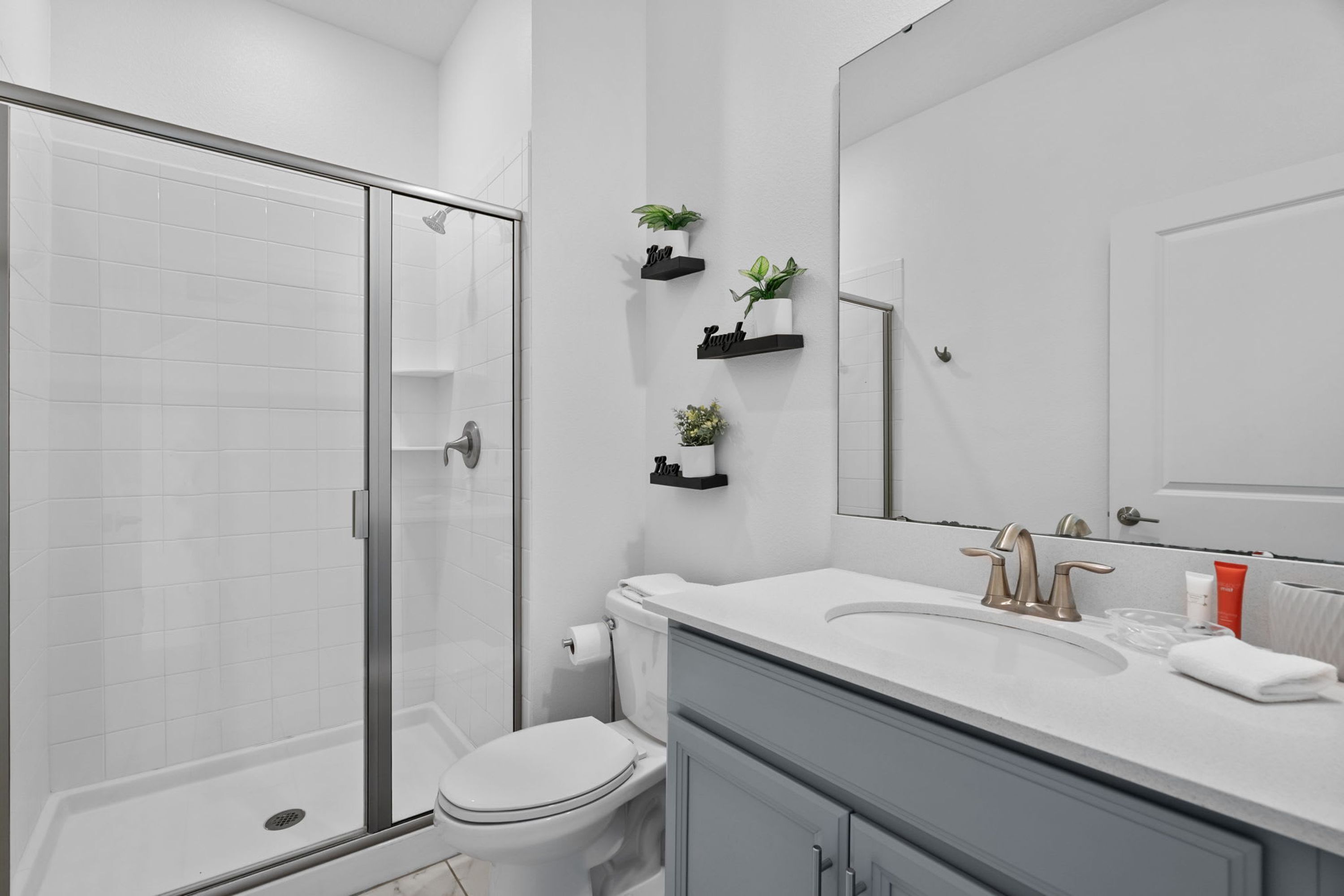 A modern bathroom with a glass shower enclosure, a white toilet, a vanity with a sink, and a mirror. There are black floating shelves with plants and a small vase.