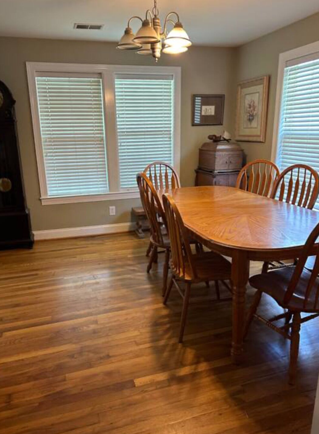 A dining area with a wooden table and chairs, large windows with blinds, a chandelier, and a cabinet.