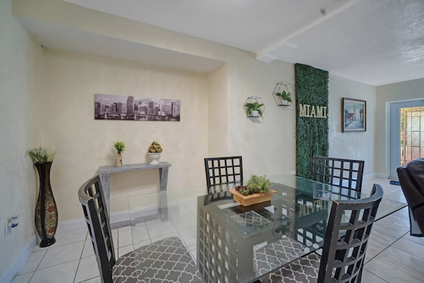Un coin repas avec une table en verre, des chaises noires, un tableau représentant un paysage urbain, un panneau indiquant « MIAMI » et une plante en pot.