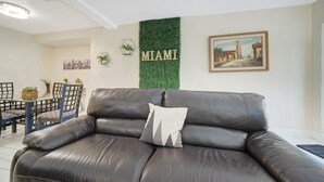 Un salon avec un canapé en cuir, une table à manger avec des chaises, un mur vert orné du mot « MIAMI » et un tableau encadré.