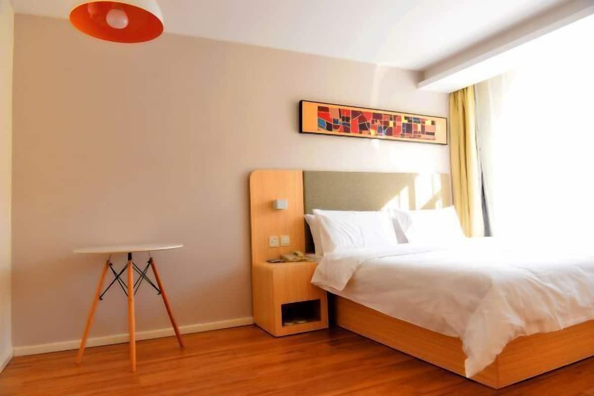 A hotel room with a bed, a bedside table, a small wooden nightstand, a wall-mounted artwork, and a window with curtains.