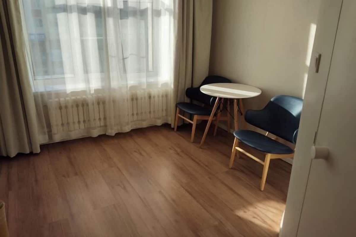 A room with a wooden floor, a small round table, two chairs, and a window with sheer curtains.