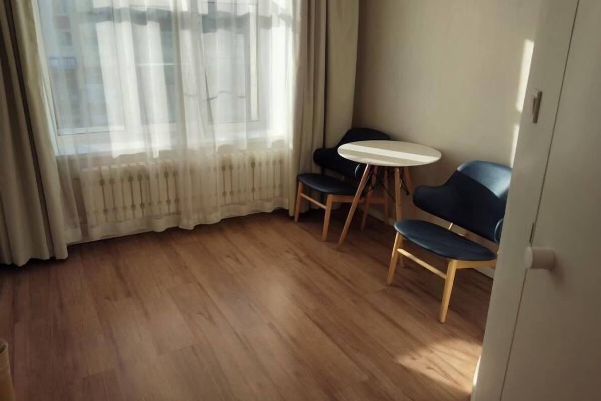 A room with a wooden floor, a small round table, two chairs, and a window with sheer curtains.