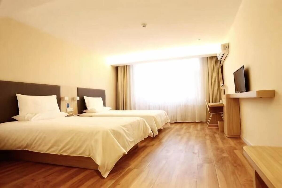A hotel room with two beds, a desk, a chair, a TV, and a large window with curtains.