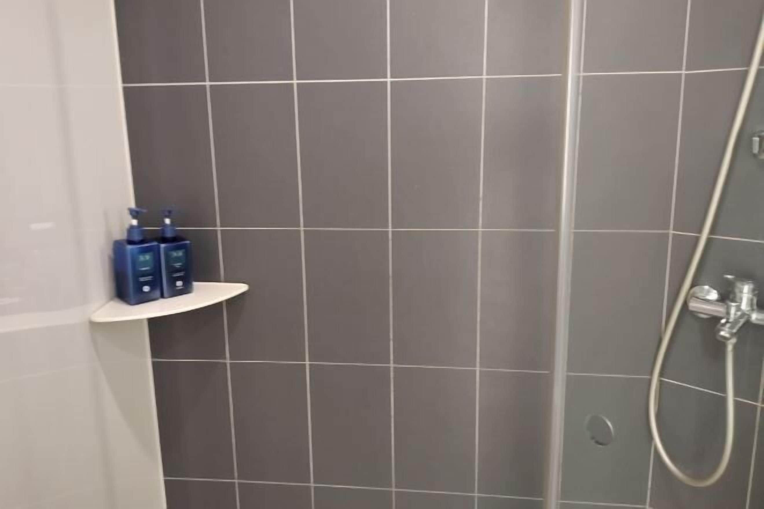 A bathroom with grey tiles, a shower, and a shelf with toiletries.