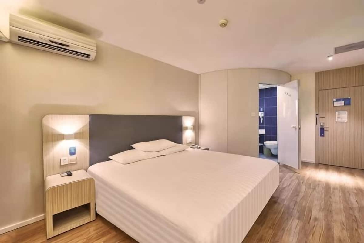 A hotel room with a large bed, bedside tables, an air conditioning unit, and a door leading to a bathroom.