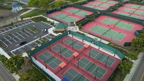 Eine Vogelperspektive einer Tennisanlage mit mehreren Plätzen, einem Parkplatz und umgebender Bepflanzung.