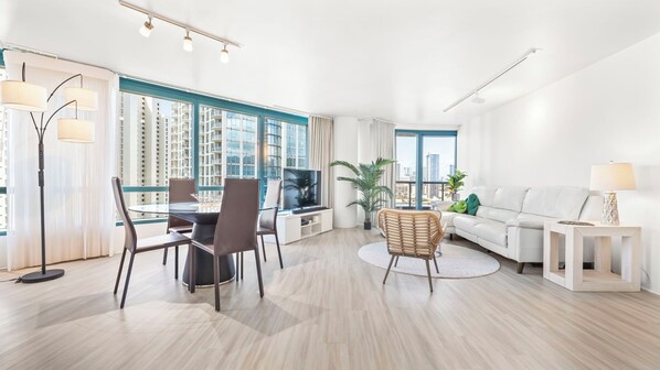 Un salon moderne comprenant un coin repas, un canapé, un téléviseur et de grandes fenêtres donnant sur la ville.
