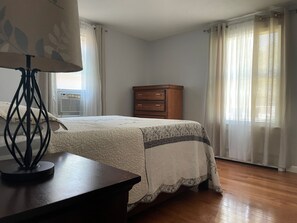 A bedroom with a bed, a wooden dresser, a lamp, and a window with curtains.