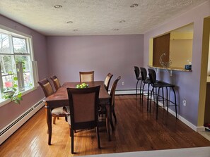 Uma área de refeições com uma mesa e cadeiras de madeira, um balcão de cozinha com banquetas e uma janela com plantas.