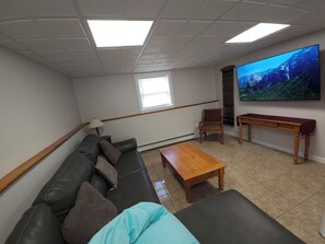 Uma sala de estar com sofá, mesa de centro de madeira, televisão fixada na parede e janela com persianas.