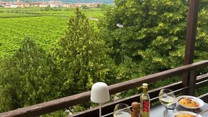 Mesa posta com pratos de massa e copos de vinho, com vista para uma paisagem verdejante com montanhas ao longe.
