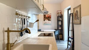 Cozinha com painel de azulejos brancos, pia e acessórios em latão. Há uma área de jantar com mesa e cadeiras. Uma estante e uma escada são visíveis ao fundo.