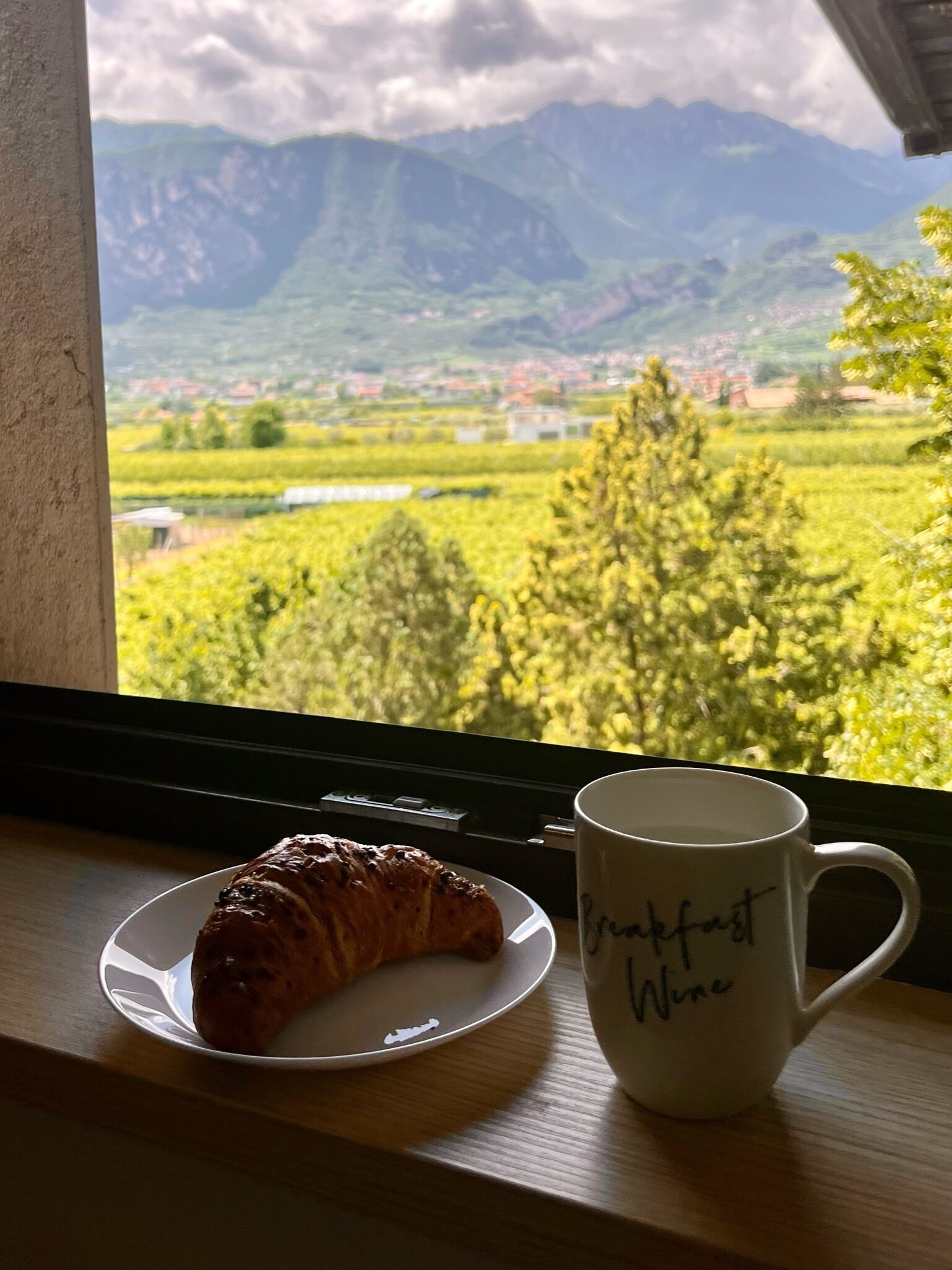 Ein Croissant auf einem Teller neben einer Tasse mit der Aufschrift 'Breakfast Wine', platziert auf einem Fensterbrett mit Blick auf einen Weinberg und Berge.