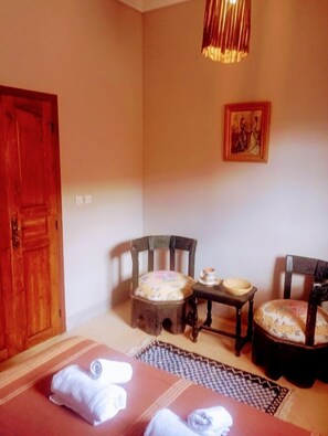 A room with a wooden door, two wooden chairs with cushions, a small table with a teapot, and a patterned rug on the floor. - Dar INNA  (Ait Benhaddou)