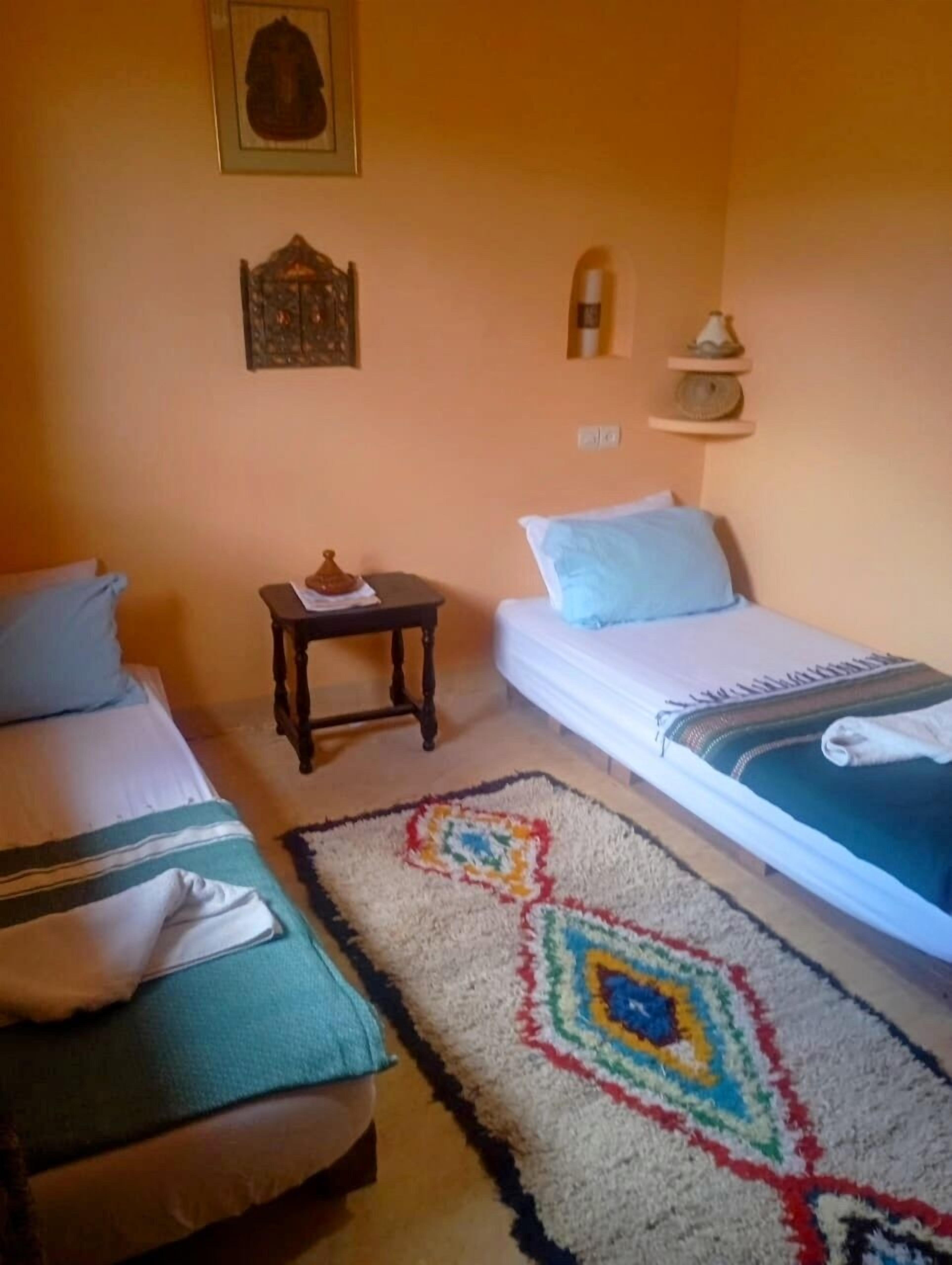 Un dormitorio con dos camas, una mesita pequeña, una estantería con adornos y una alfombra estampada.