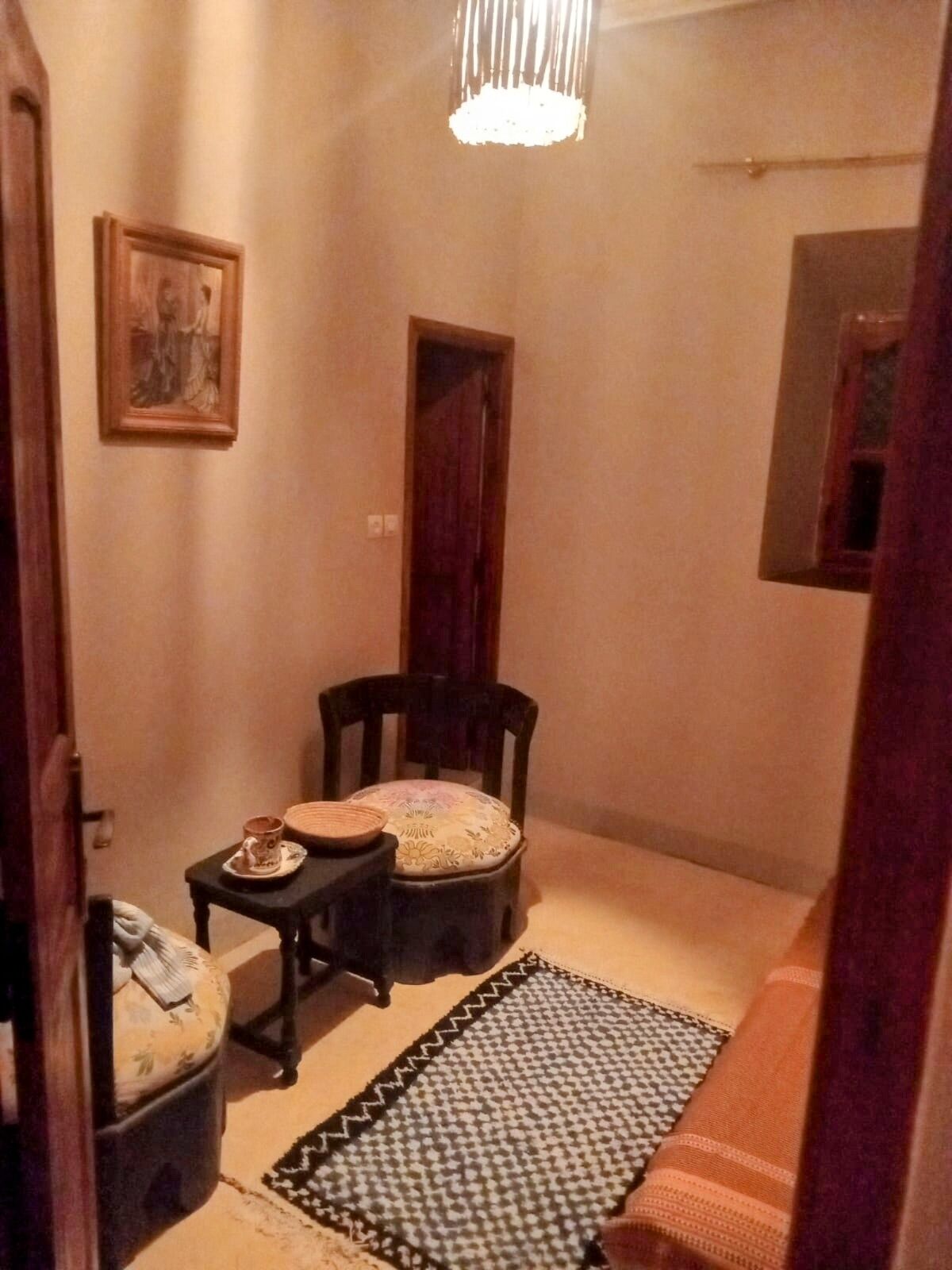 A small, cozy room with a wooden chair, a small table with a cup and saucer, a patterned rug, and a framed picture on the wall.