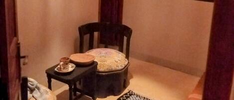 A small, cozy room with a wooden chair, a small table with a cup and saucer, a patterned rug, and a framed picture on the wall.