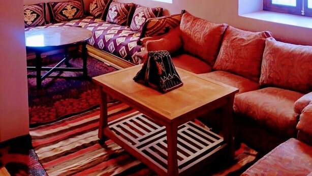 A cozy living room with a sofa, a coffee table, and a patterned rug. There are two windows with curtains, a wall-mounted lamp, and a decorative plate. - Dar INNA  (Ait Benhaddou)